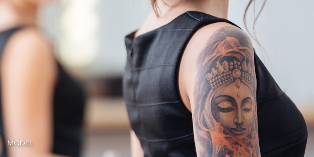 Back view of pretty young woman in black dress with tattoo on her hand standing in front of the mirror Back view of pretty young woman in black dress with tattoo on her hand standing in front of the mirror